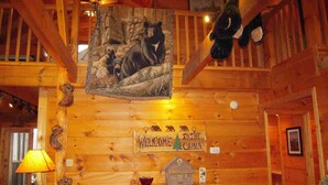 Interior de una cabaña con paredes de madera, un cuadro de un oso, un letrero de bienvenida, una lámpara y una pequeña maqueta de una casa.