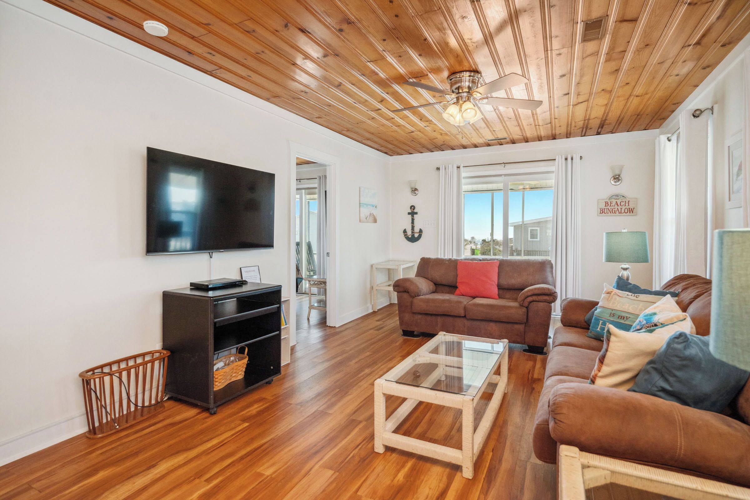 Ein Wohnzimmer mit Holzbalkendecke, einem Fernseher, einem braunen Sofa, einem Glastisch und einem Schild mit der Aufschrift 'BEACH BUNGALOW'.