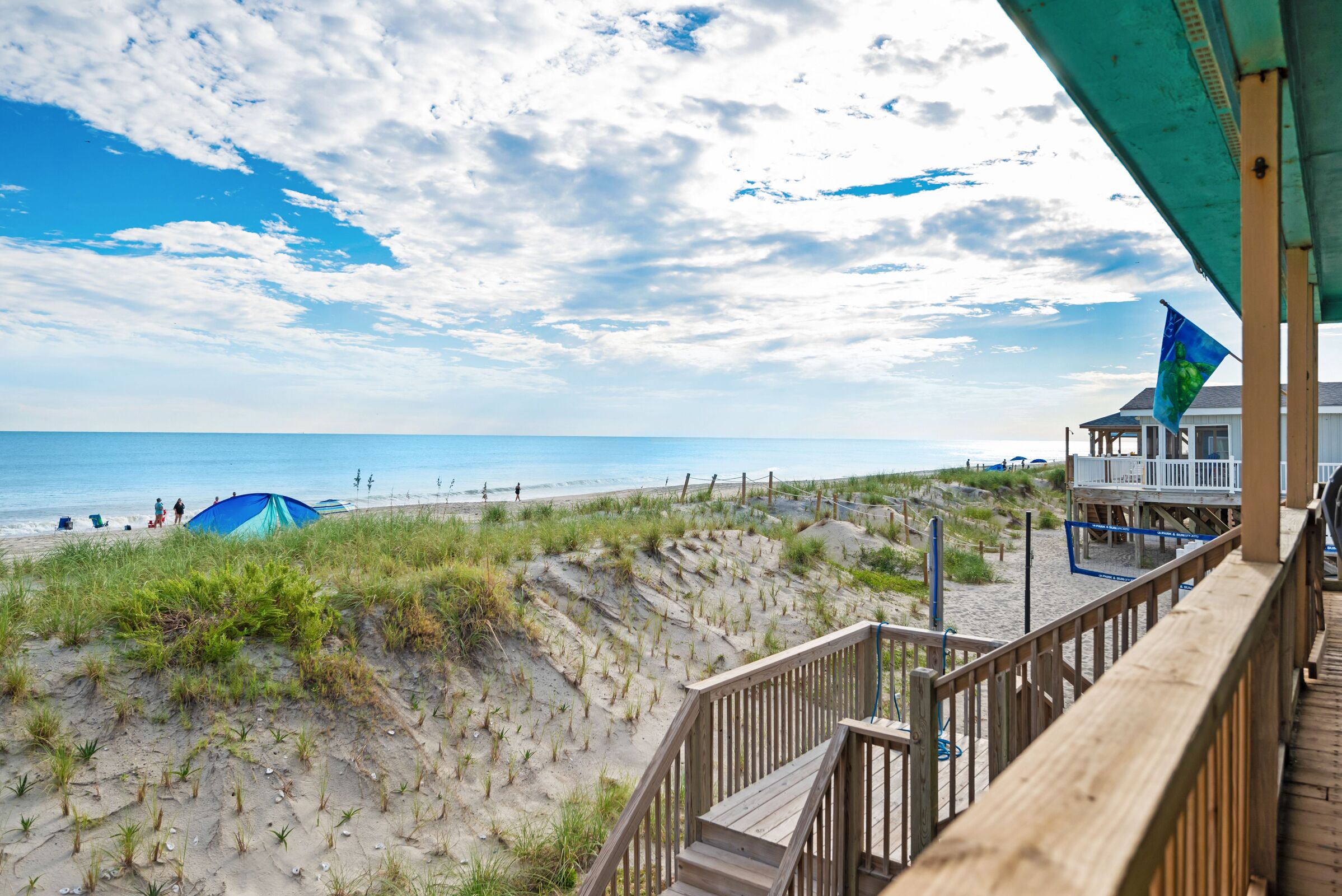 Une vue sur la plage avec une terrasse en bois, une tente bleue et un bâtiment surmonté d’un drapeau.