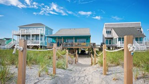 A beachfront property with a wooden walkway leading to a house with a balcony. - A Pirate's Paradise: Oceanfront Home with Covered Porch and Porch Swing (Oak Island)