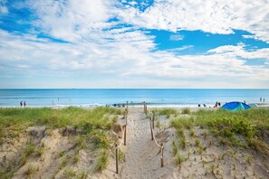 Un sentier de sable qui longe la plage et mène à l’océan, avec une tente et des personnes visibles au loin.
