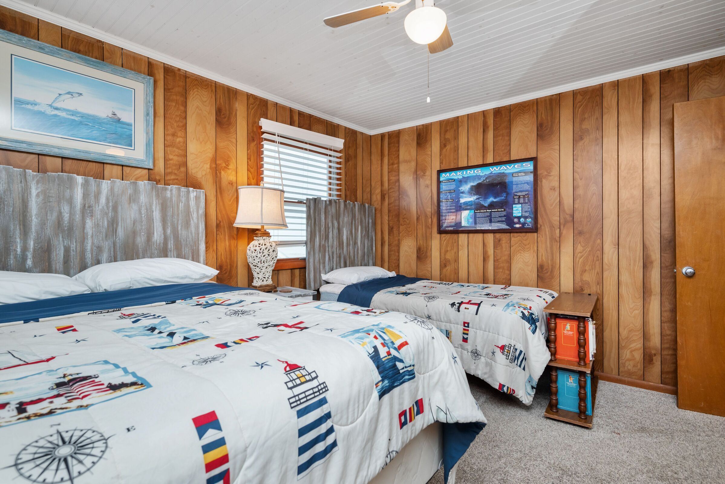 A room with wooden paneling, two beds with nautical-themed bedding, a ceiling fan, a framed picture of a whale, and a poster on the wall.