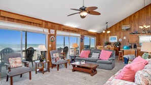 A spacious living room with a large window overlooking the ocean, wooden paneling, and a ceiling fan. - A Pirate's Paradise: Oceanfront Home with Covered Porch and Porch Swing (Oak Island)