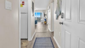 Un couloir avec une porte blanche, un réfrigérateur, un tapis bleu et une vue sur un coin repas avec une table et des chaises.