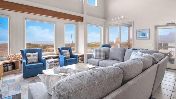Un salón amplio con un sofá seccional gris grande, un sillón azul y una mesa de centro blanca. Varias ventanas ofrecen vistas del paisaje.