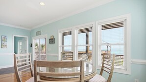 A dining area with a round glass table and wooden chairs, white-framed windows, and a view of the ocean.