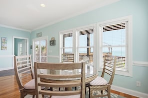 A dining area with a round glass table and wooden chairs, white-framed windows, and a view of the ocean.