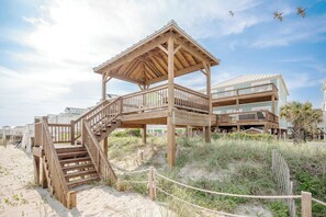 Une maison de plage en bois dotée d’un balcon, d’un escalier menant au bâtiment principal et d’une plage de sable.