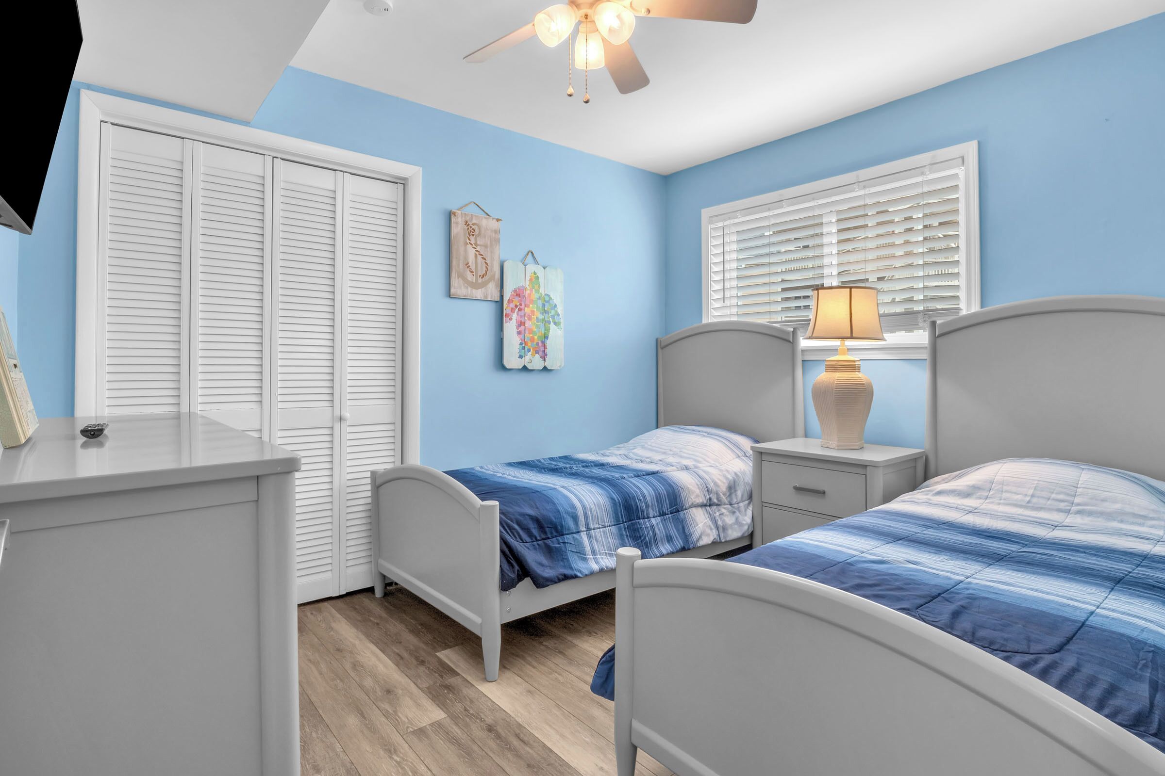 A bedroom with two beds, a nightstand, a dresser, a ceiling fan, and a window with blinds.