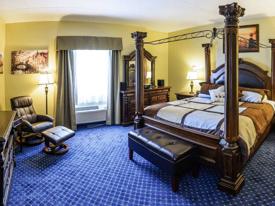 A four-poster bed with a wooden headboard, a leather armchair, a bench, and a desk with a lamp.