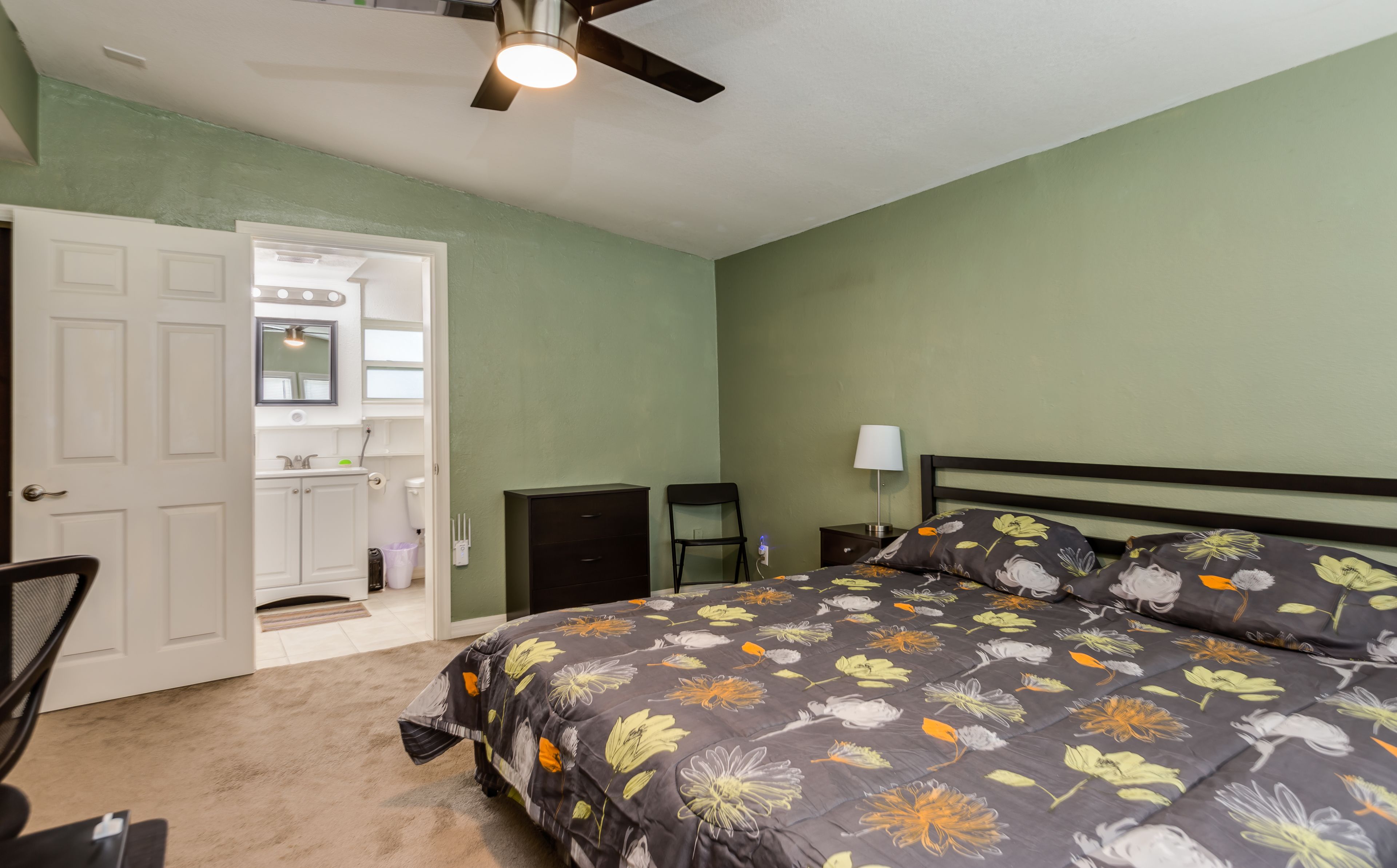 A bedroom with a bed, a ceiling fan, a nightstand, a chair, and a door leading to a bathroom.