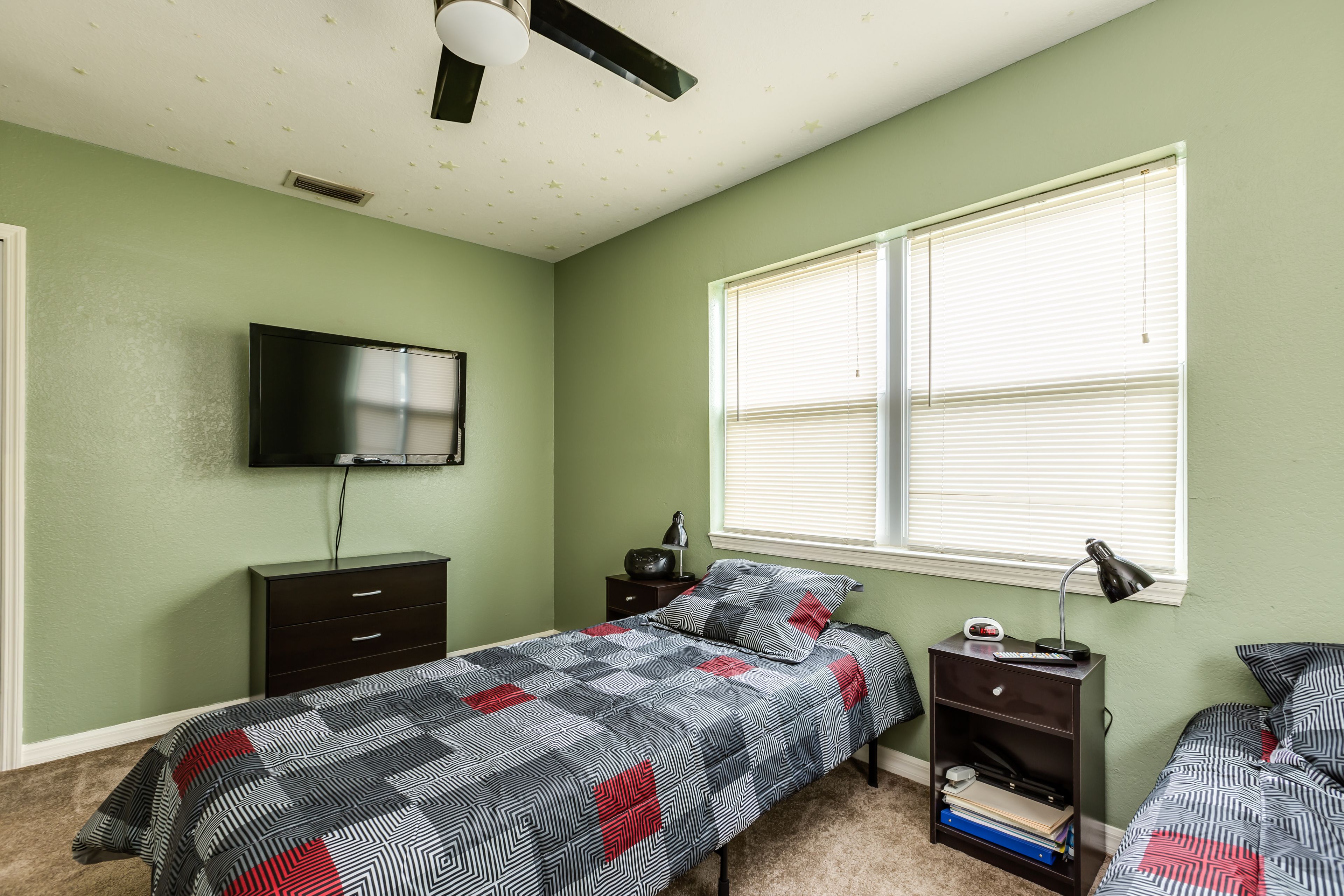A bedroom with a bed, a nightstand, a dresser, a TV mounted on the wall, and a window with blinds.