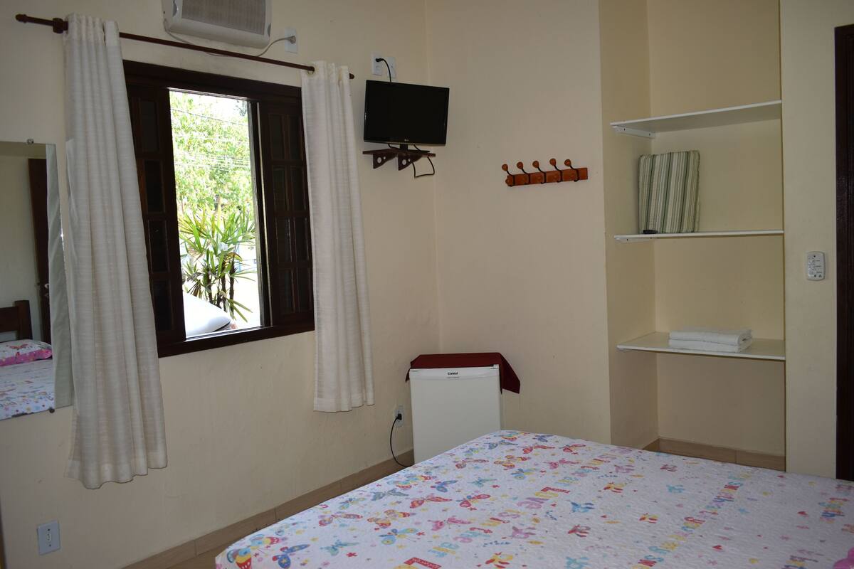Une chambre avec un lit, une télévision, une fenêtre avec des rideaux, un petit réfrigérateur et une étagère avec des serviettes.