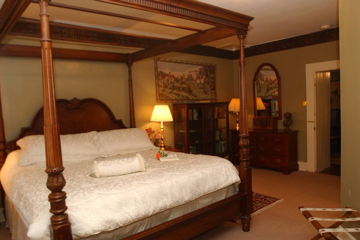 A four-poster bed with a white bedspread and pillows, a wooden headboard, and a nightstand with a lamp. There is a wooden dresser with a mirror and a painting on the wall.
