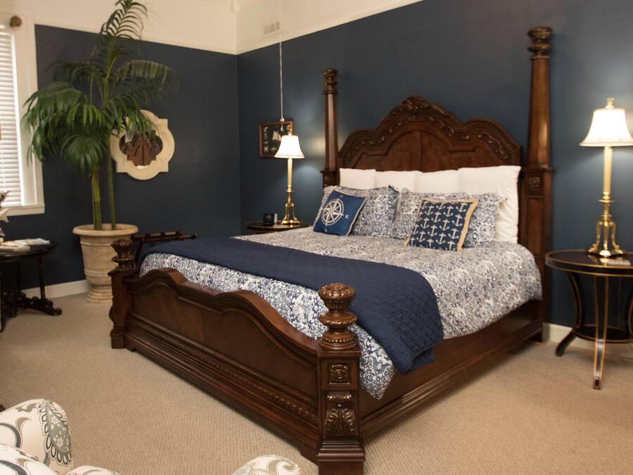 A bedroom with a large bed, two bedside tables, a nightstand, a potted plant, and a window with blinds.