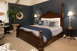 A bedroom with a large bed, two bedside tables, a nightstand, a potted plant, and a window with blinds. - Jailer's Inn (Bardstown)