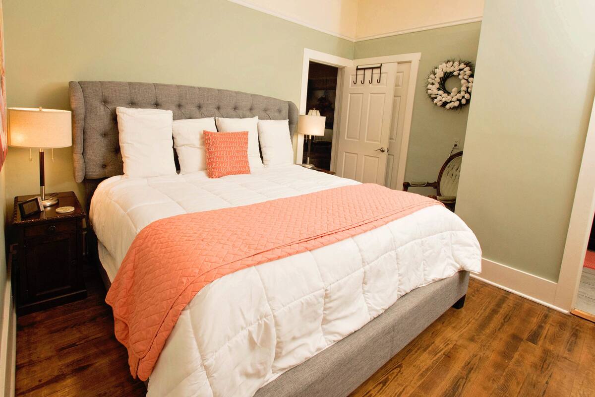 A neatly made bed with a grey headboard, white and orange pillows, and a white quilt. There is a wooden nightstand with a lamp beside the bed.