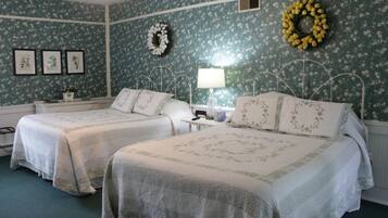A room with two beds, floral wallpaper, a nightstand with a lamp, and a wreath on the wall.