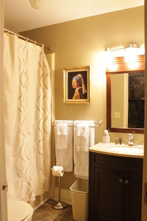 A bathroom with a shower curtain, a toilet, a sink with a mirror, and a framed picture on the wall. - Jailer's Inn (Bardstown)