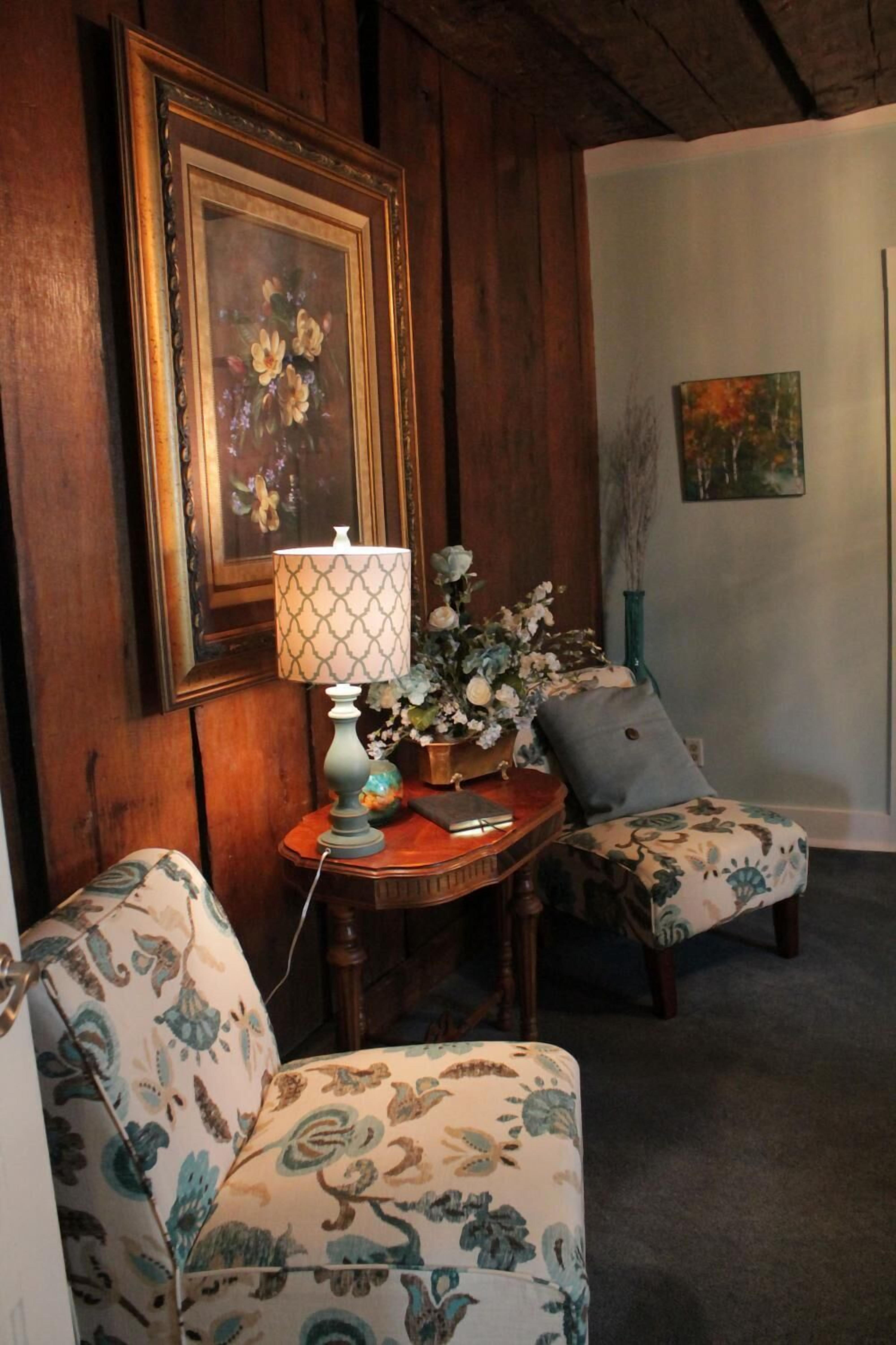 A cozy room with wooden paneling, a framed floral painting, a table with a lamp, and two patterned armchairs.