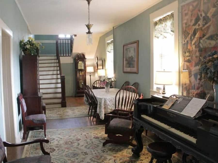 A spacious living room with a grand piano, a dining table, and a staircase leading to an upper floor.