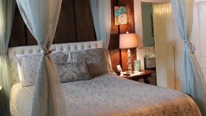 A bedroom with a canopy bed, a bedside table with a lamp, and a door leading to another room.
