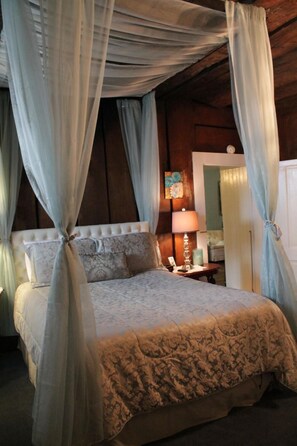 A bedroom with a canopy bed, a bedside table with a lamp, and a door leading to another room.
