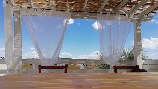 A covered outdoor seating area with a wooden table and chairs, a pergola with white curtains, and a view of the countryside. - B&b Villa Kairos (Ioppolo Giancaxio)