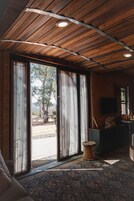 Una habitación con techo de madera, grandes puertas correderas de vidrio que dan a un espacio al aire libre, un taburete tejido y un televisor de pantalla plana sobre un soporte.