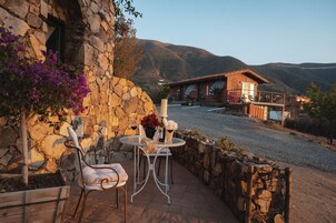 Edificio de piedra con un patio que incluye dos sillas blancas y una mesa, un muro de piedra y vistas a una montaña.