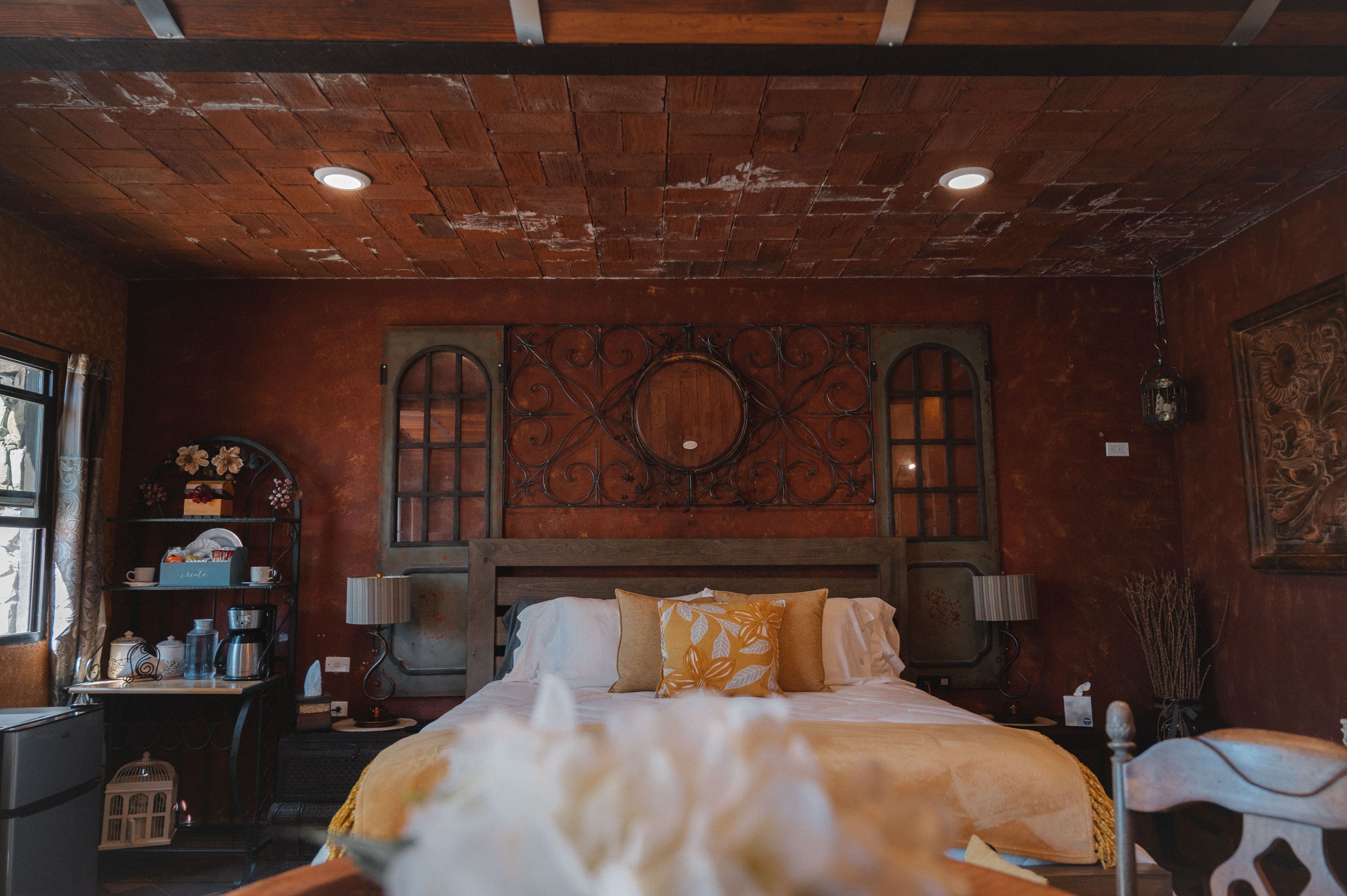 Una habitación con techo de madera, una cama con ropa de cama blanca y amarilla, un escritorio con una lámpara y un espejo decorativo sobre la cama.