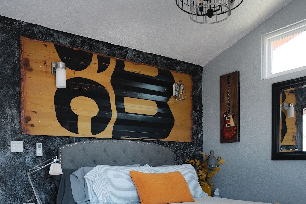 Quarto com uma cama grande, uma mesa de cabeceira, uma luminária, um objeto decorativo na parede e um violão.