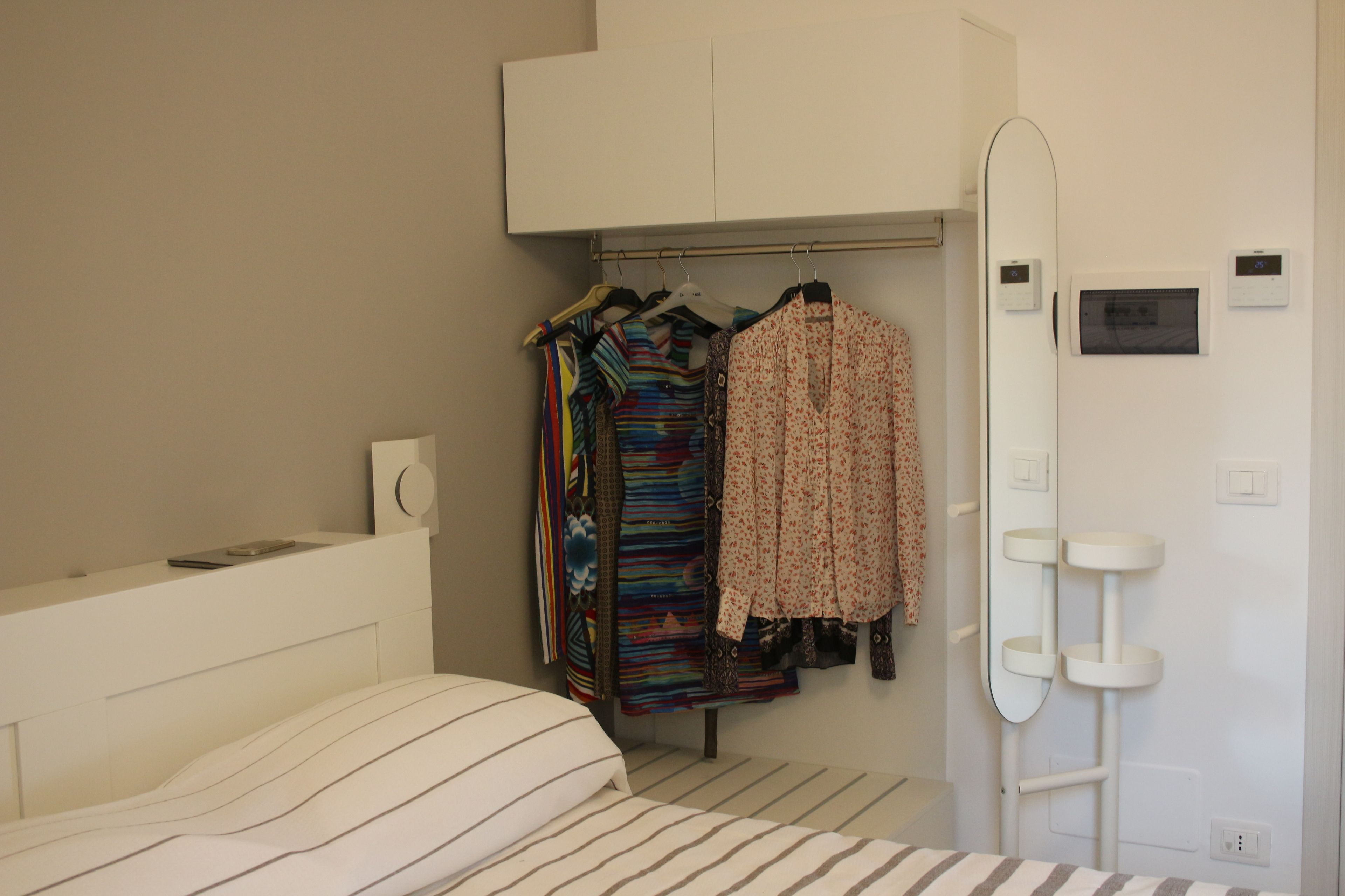 Un dormitorio con cama, un armario con ropa colgada, un espejo y un mueble blanco sobre el armario.
