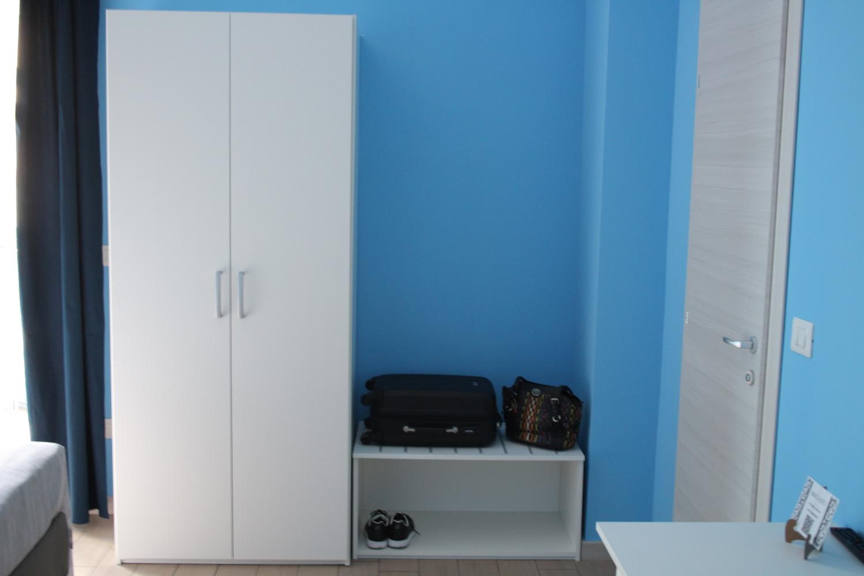 Una habitación con un armario blanco, un banco con una maleta y un bolso, y un par de zapatos en el suelo.