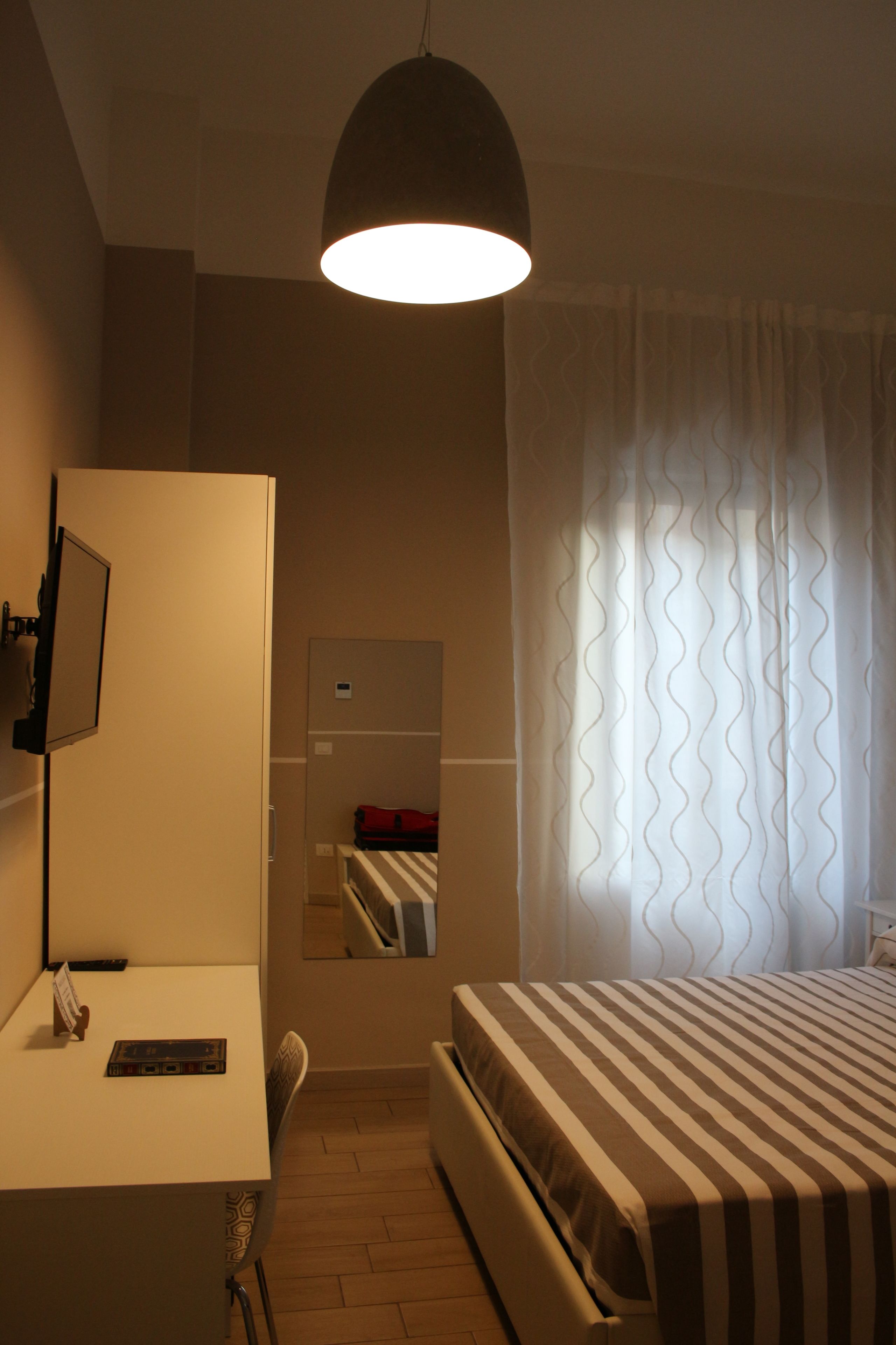 Habitación de hotel con cama, escritorio, silla, televisor, espejo y ventana con cortinas.
