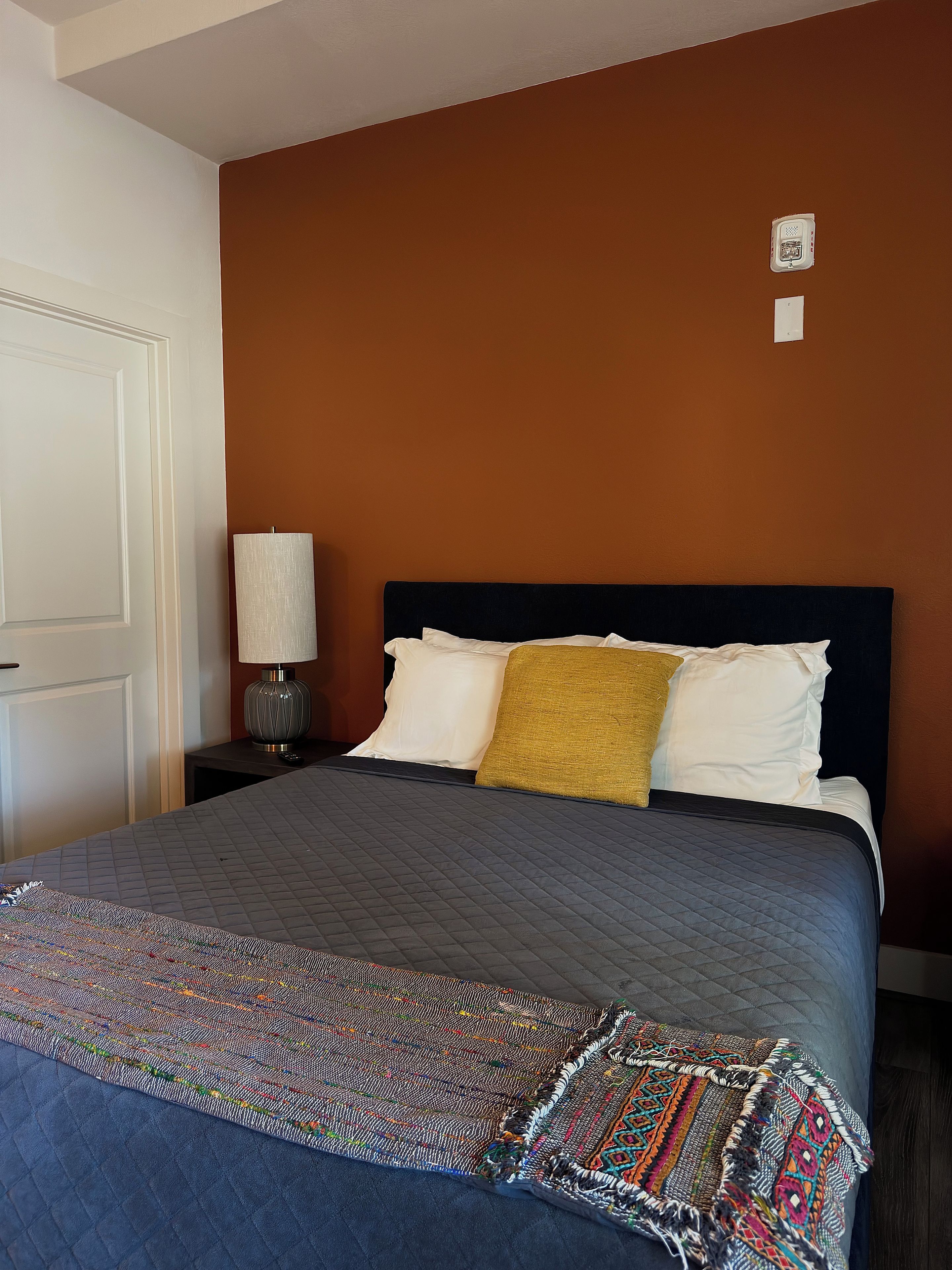 Una cama bien hecha con cubierta acolchada, una lámpara de noche y un interruptor de luz en la pared.