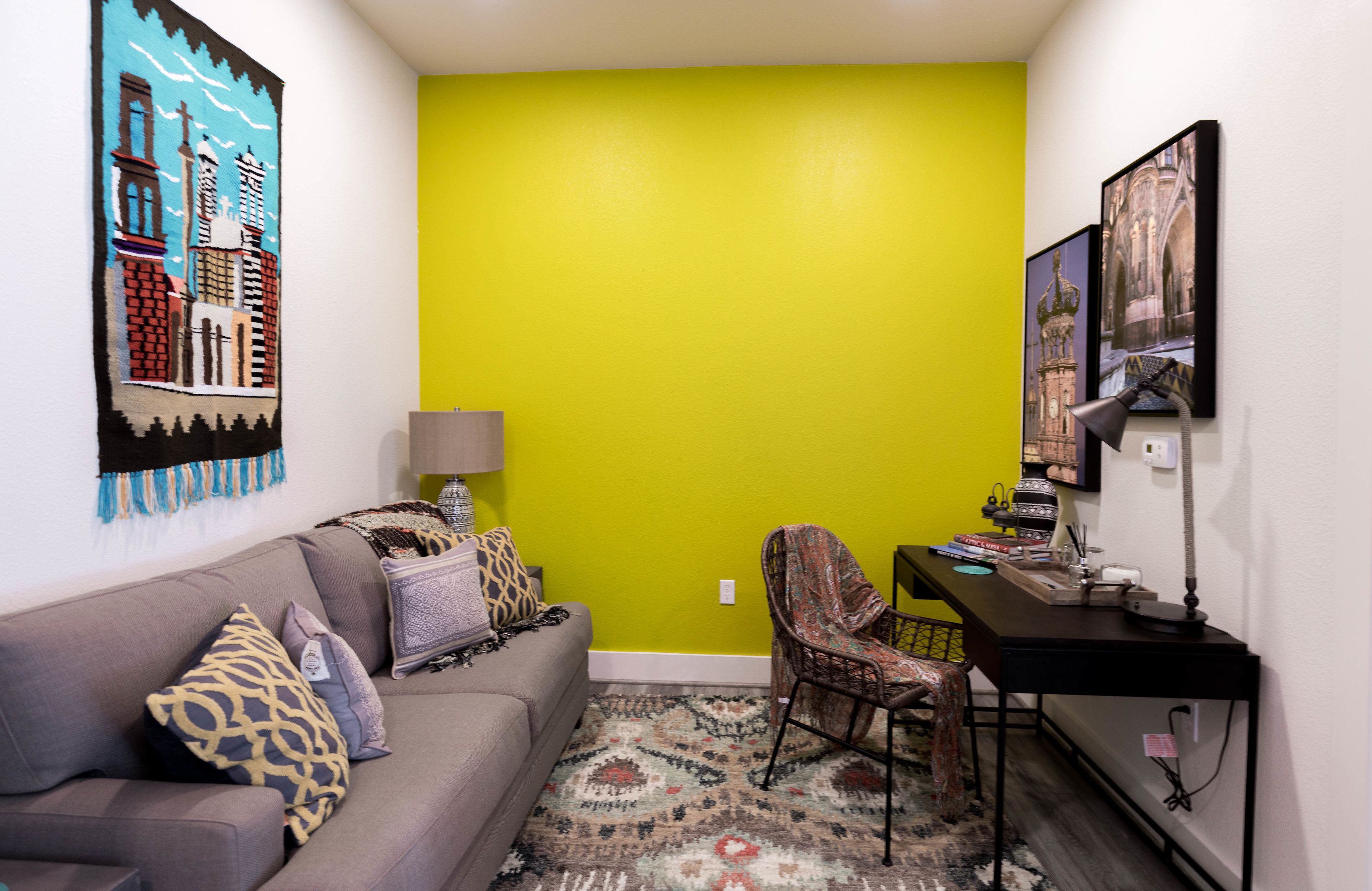 Una sala de estar con una pared verde de acento, un sofá gris con cojines estampados, un tapete con diseño, un sillón de mimbre y un escritorio con una lámpara y libros.