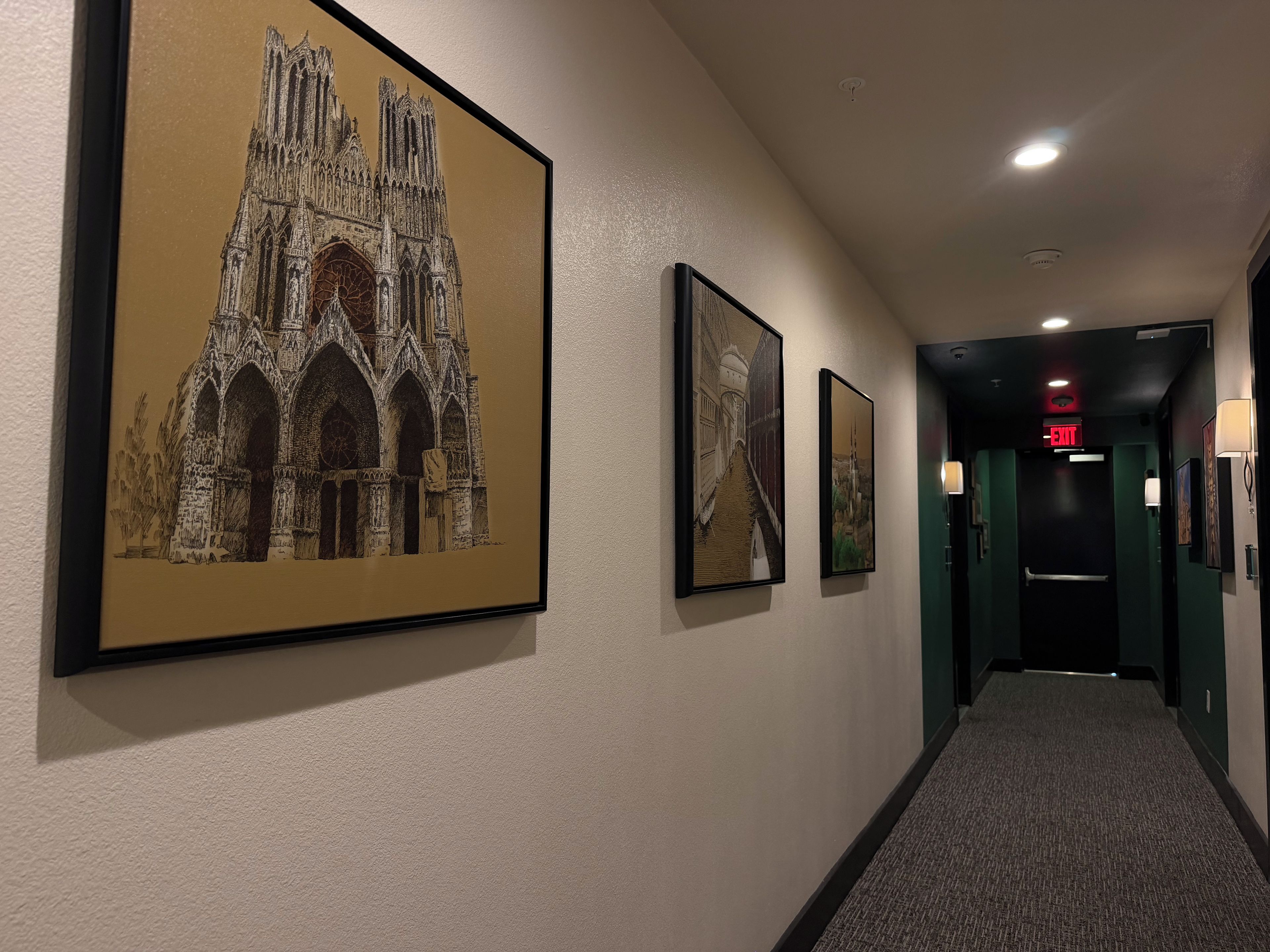 A hallway with framed pictures of Gothic architecture, a carpeted floor, and an exit sign.
