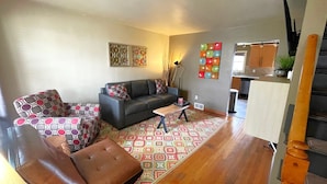 A living room with a sofa, ottoman, and coffee table, a kitchen area with cabinets and a sink, and a staircase leading to another level.