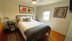 A neatly arranged bedroom with a bed, bedside tables, a nightstand, a floor lamp, a window, and artwork on the walls. - Escape to our cozy retreat, just a 10-min drive to Greensburg (Youngwood)