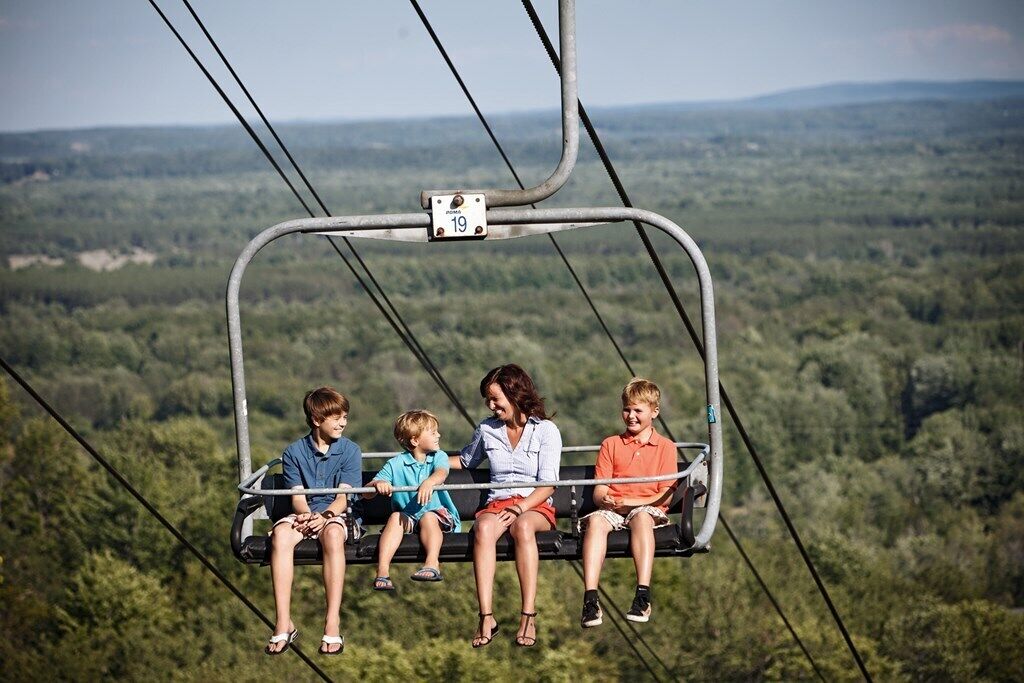 Eine Seilbahn mit vier Personen, darunter zwei Kindern und einem Erwachsenen, fährt über eine bewaldete Landschaft.