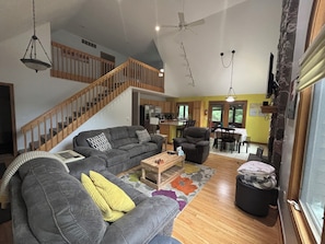 A spacious living area with a gray sectional sofa, a wooden coffee table, and a dining table with chairs. A staircase with wooden railings leads to an upper level.