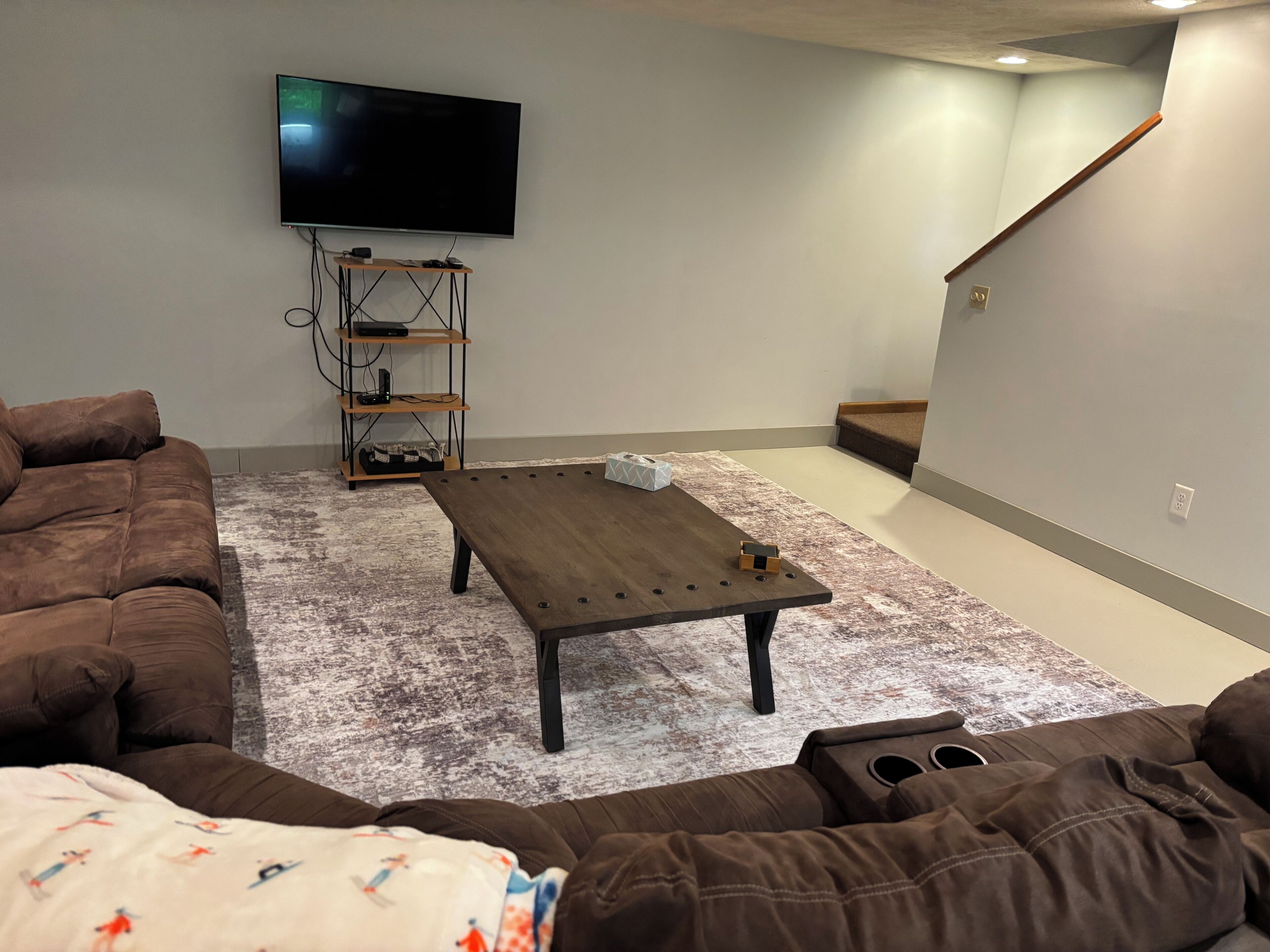 A living room with a brown couch, a coffee table, a TV mounted on the wall, and a staircase in the background.