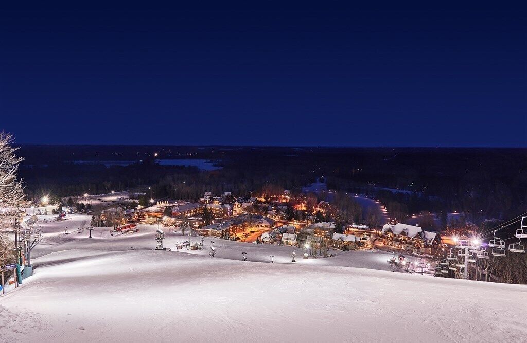 A snow-covered ski resort at night with illuminated buildings and ski lifts.