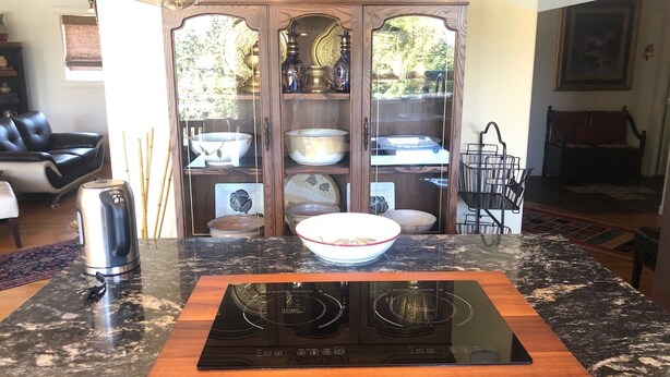 Una cocina con tope de inducción negro, encimera de madera y un armario de vidrio con vajilla.