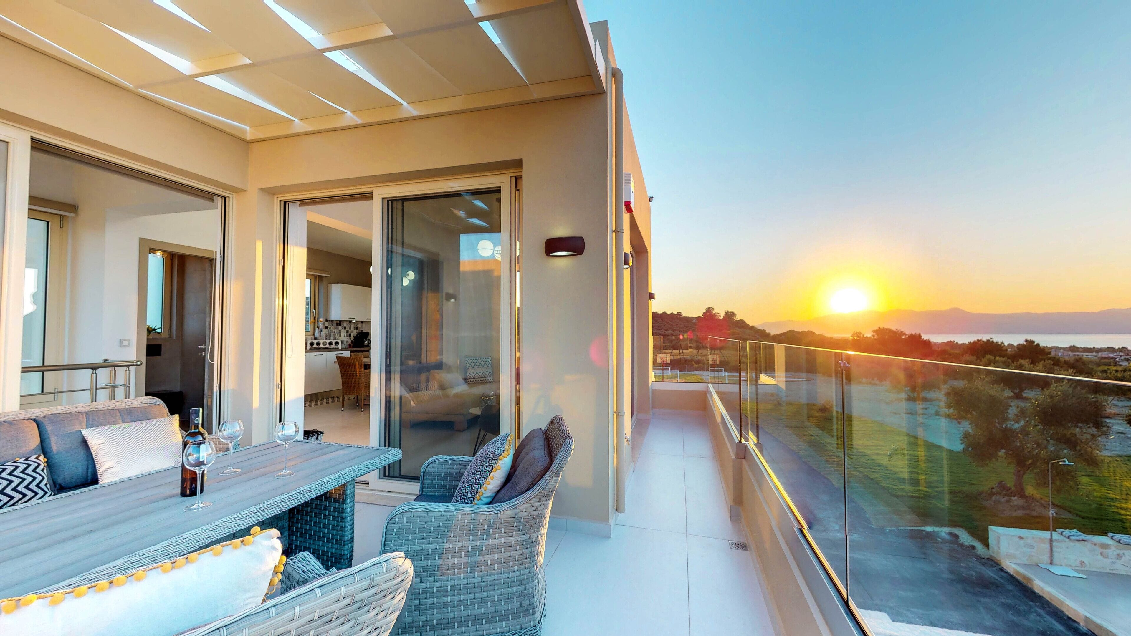 Een balkon met een eethoek, een glazen deur naar een keuken, en uitzicht op de ondergaande zon boven de zee.