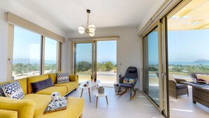 A bright living room with a yellow sofa, a rocking chair, and a dining table. Large windows offer a view of the landscape. - LIBERTY &  FREEDOM villa, helipad and pools!Perfect for large groups up to 16 ! (Platanias)