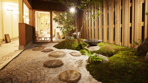 A well-lit outdoor area with a stone pathway leading to a wooden fence, surrounded by greenery and moss. - utsuwa Designed Hostel (Kyoto)