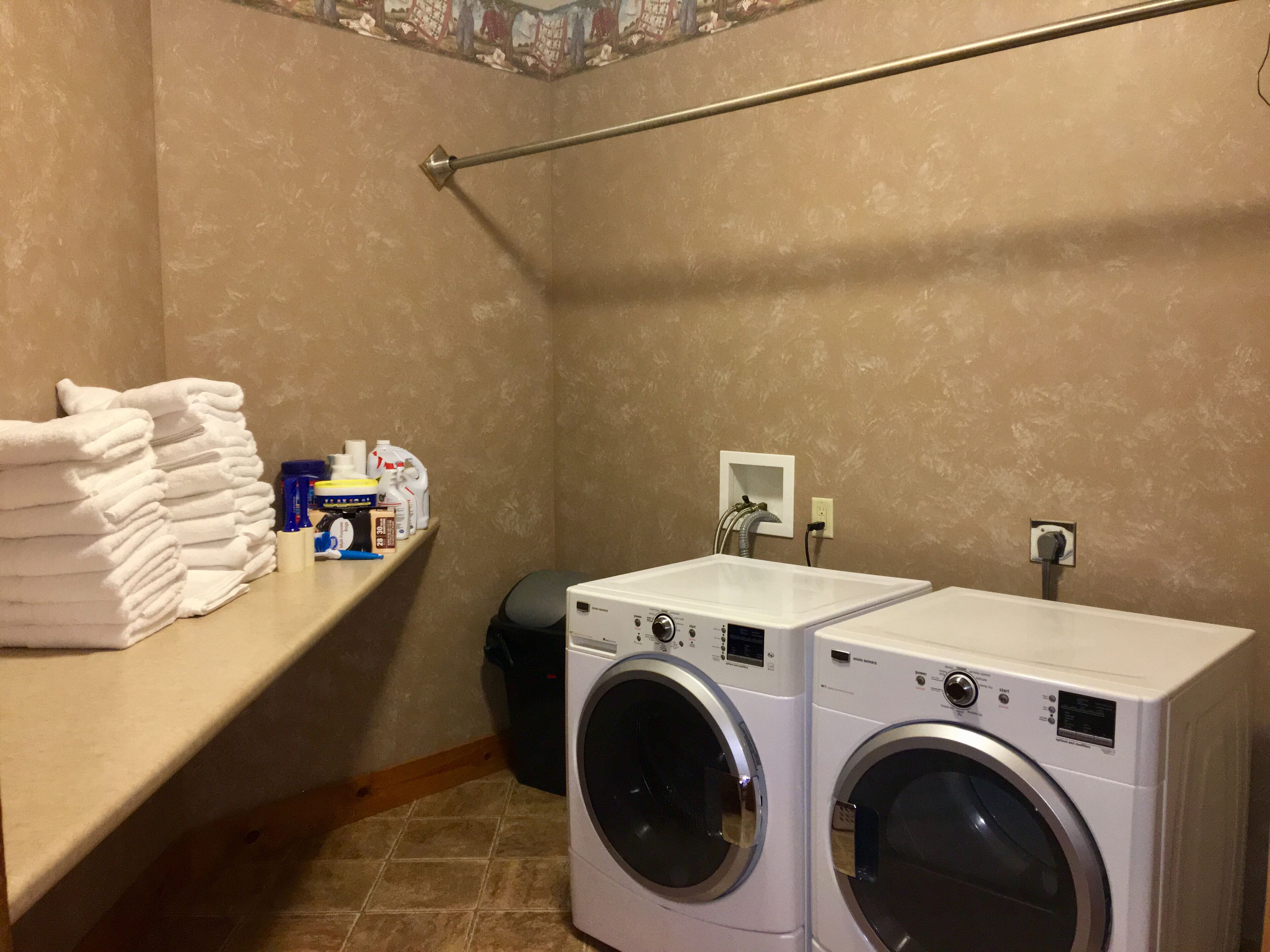 A laundry room with two stacked washing machines, a shelf with folded towels, and cleaning supplies.