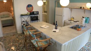A compact kitchen with a central island, a microwave, a refrigerator, and a sink. There are bar stools along the island and a small seating area with a chair and a table. - Oleander - Charming Cottage Downtown - Steps to the Beach! (Flagler Beach)